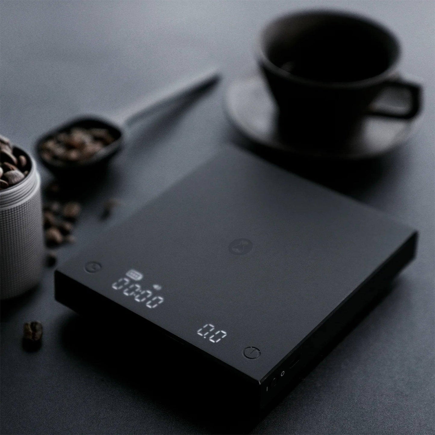 Die Blackmirror Basic Plus Kaffeewaage in Schwarz mit Kaffeebohnen und einer Tasse auf dem Tisch.